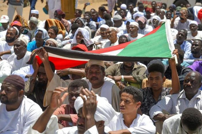 Sudanese protesters use a national flag to shield themselves from sun as they attend Friday prayers near the military headquarters