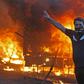 A protester flashes the V for victory sign as a riot police vehicle burns behind him during clashes amidst demonstrations against state corruption, failing public services, and unemployment in the Iraqi capital