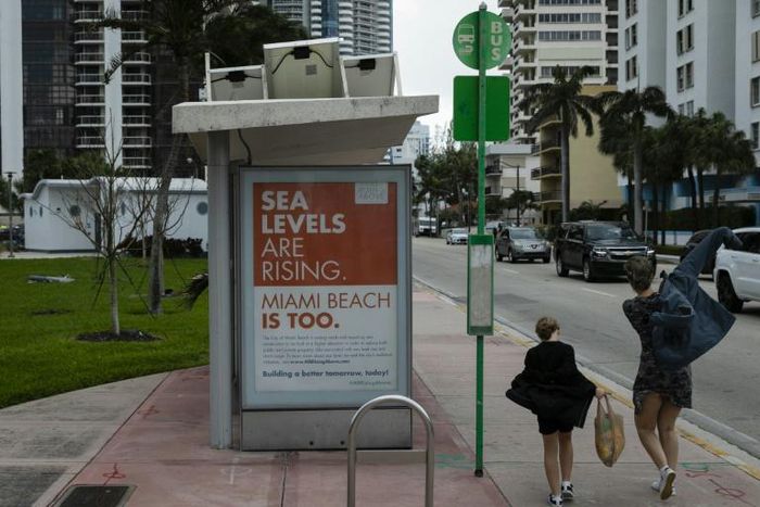 People walk past a billboard in Miami Beach discussing sea level rise: dozens of trucks have started dumping hundreds of thousands of tons of sand on Miami Beach as part of US government measures to protect Florida's tourist destinations against the ef...