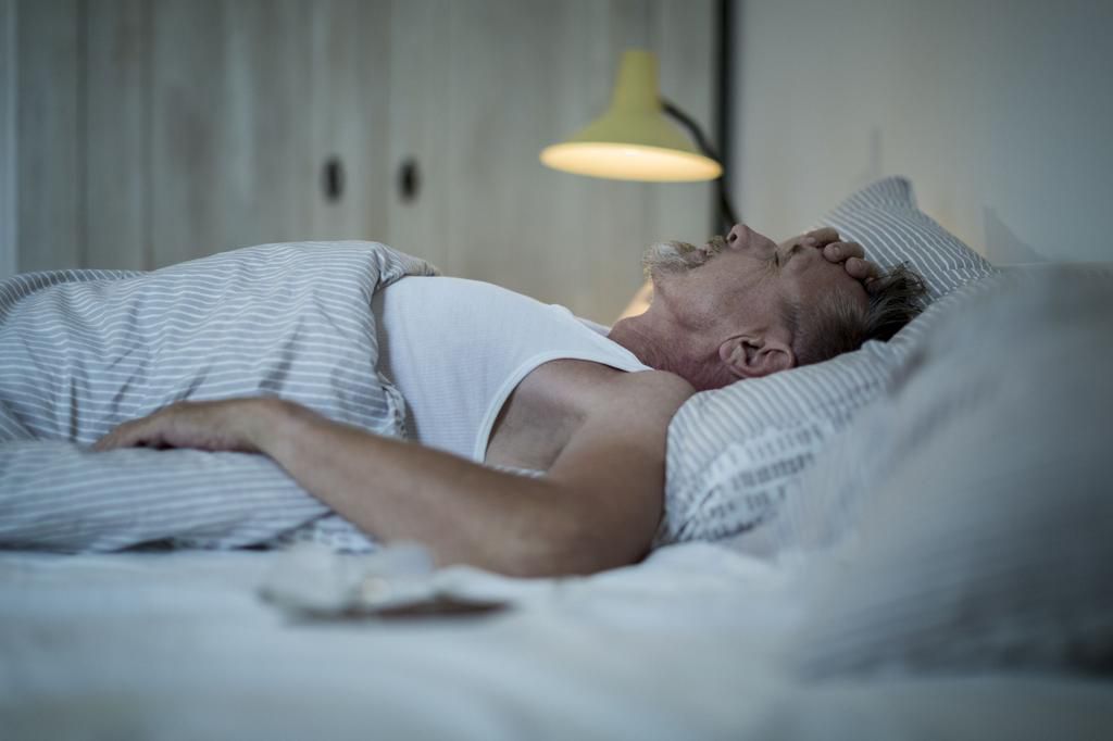 Senior man in his early 60s with greying beard is sick and sleepless in bed while night.