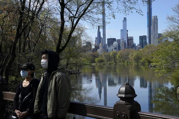 The Midtown Manhattan skyline is reflected in the water as people in masks walk along the lake in Central Park