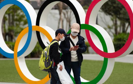 People in Japan wear masks due to the COVID-19 pandemic in front of Olympics rings.