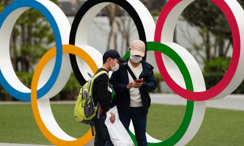 People in Japan wear masks due to the COVID-19 pandemic in front of Olympics rings.
