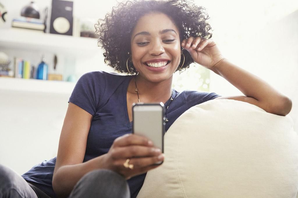 Smiling African American woman texting with cell phone on living room sofa