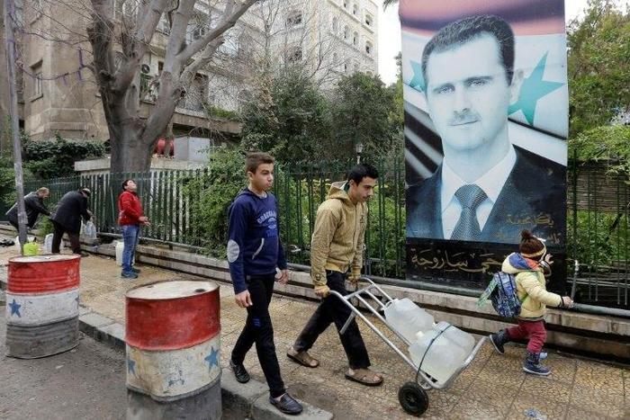 Syrians walk past a poster of President Bashar al-Assad as they go to fill plastic containers with water at a public fountain in the capital Damascus