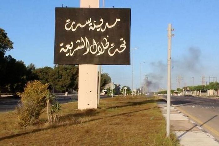 A sign which reads in Arabic, "The city of Sirte, under the shadow of Sharia" is seen as smoke rises in the background while forces aligned with Libya's new unity government advance on the eastern and southern outskirts of the Islamic State stronghold ...