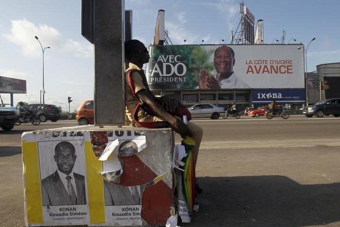 A student sits next to campaign posters of independent candidate Kouadio Konan Bertin, with a campaign billboard Ivory Coast President Alassane Ouattara seen in the background, ahead of the October 25 presidential election, in Abidjan October 21, 2015....