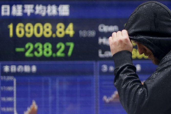 A pedestrian taking shelter from rain walks past an electronic board showing the Japan's Nikkei average outside a brokerage in Tokyo, Japan, January 18, 2016. REUTERS/Yuya Shino
