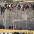 African migrants sit atop a border fence as Spanish police in riot gear try to turn