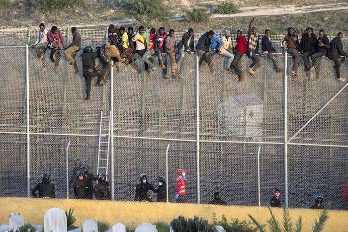 African migrants sit atop a border fence as Spanish police in riot gear try to turn