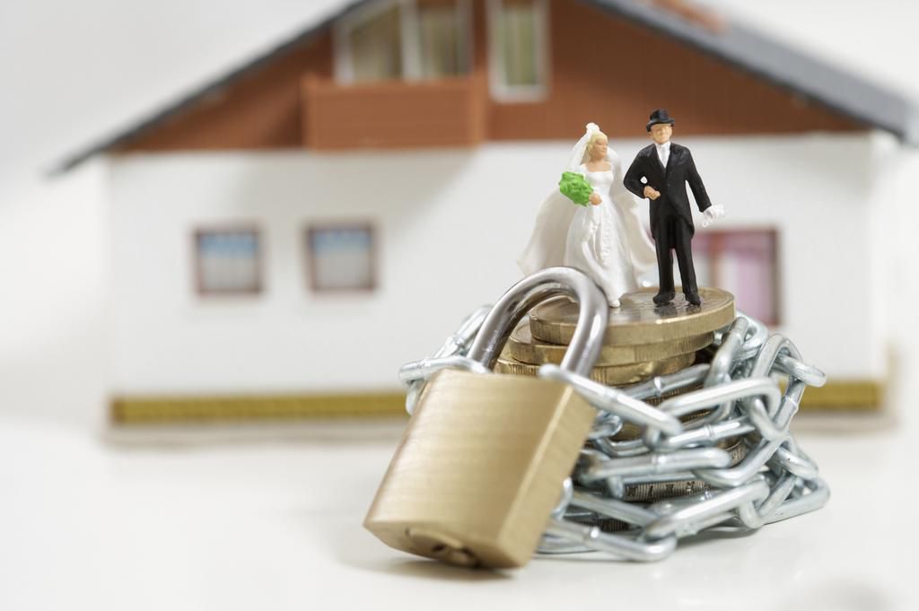 Bridal couple figurines on a stack of coins wrapped of a chain with padlock, miniature house in background
