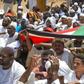 Sudanese protesters use a national flag to shield themselves from sun as they attend Friday prayers near the military headquarters