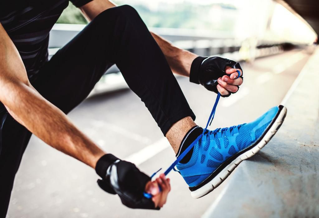 Unrecognizable young sporty man with fingerless gloves tying shoelaces outdoors in the city.
