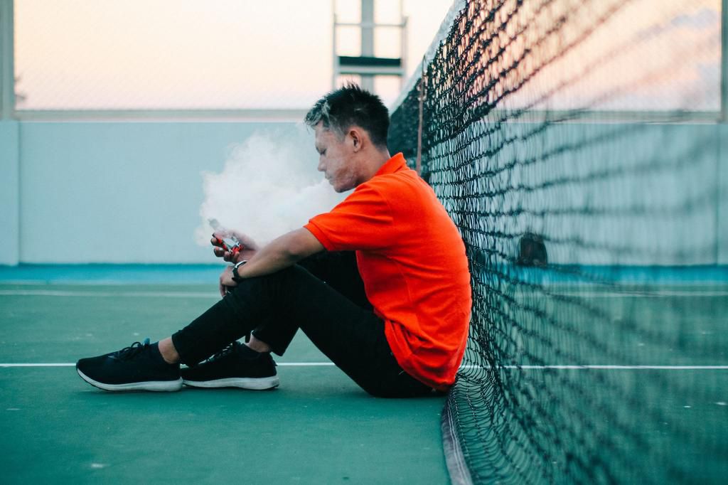 Side View Of Young Man Smoking Electronic Cigarette While Sitting On Tennis Court