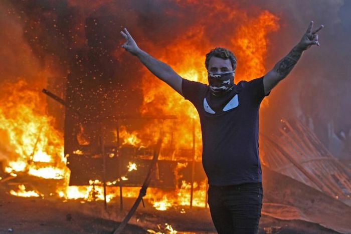 A protester flashes the V for victory sign as a riot police vehicle burns behind him during clashes amidst demonstrations against state corruption, failing public services, and unemployment in the Iraqi capital
