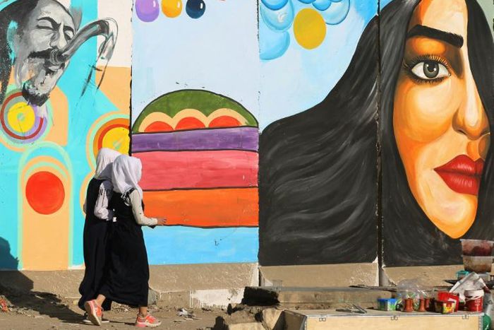 Iraqi women walk past a mural inspired by ongoing anti-government protests in Tahrir square in Baghdad while political leaders are struggling to choose a new prime minister