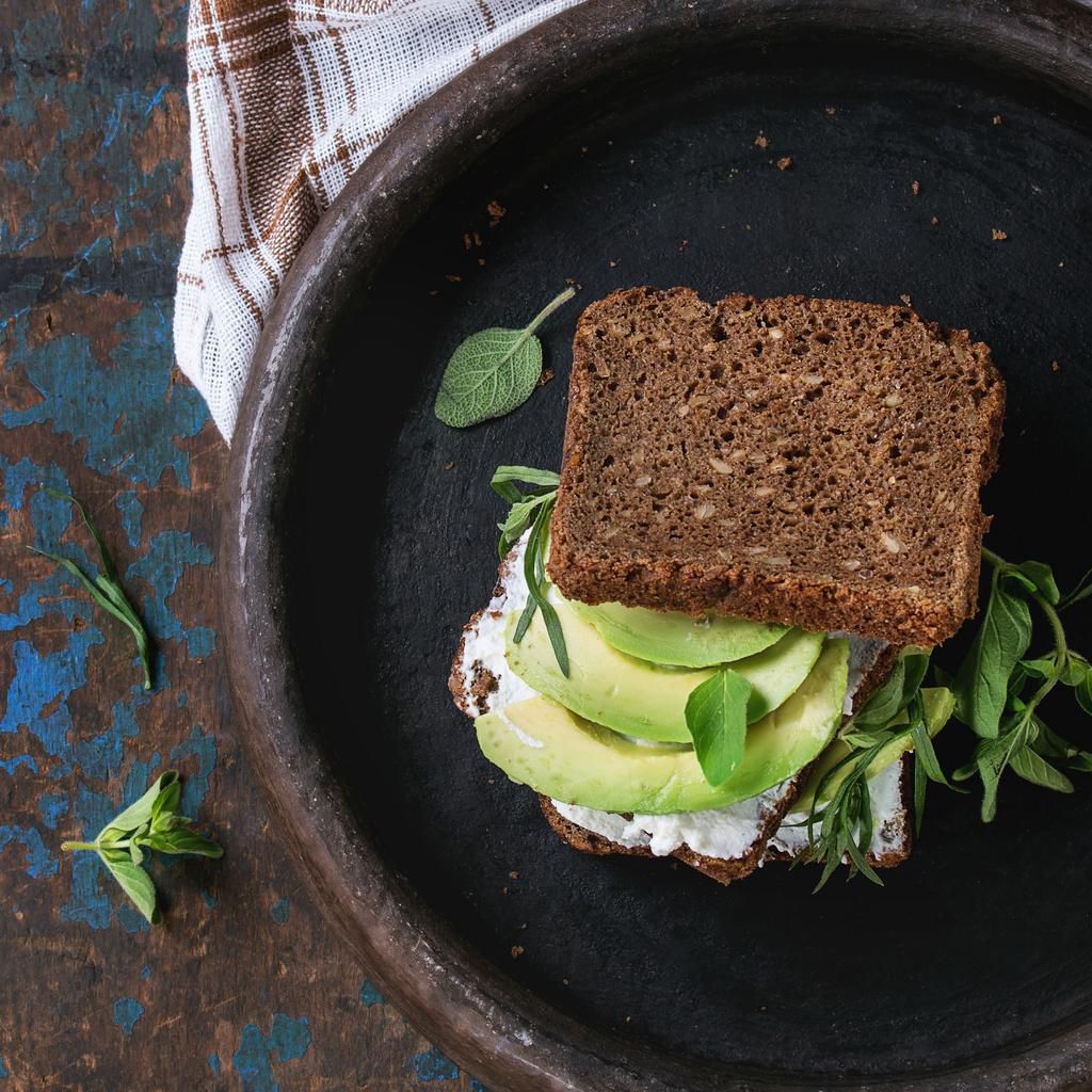 Avocado and ricotta rye sandwich with fresh herds on black clay tray with checkered kitchen towel over dark wooden textured background. Healthy eating theme. Top view. Square image