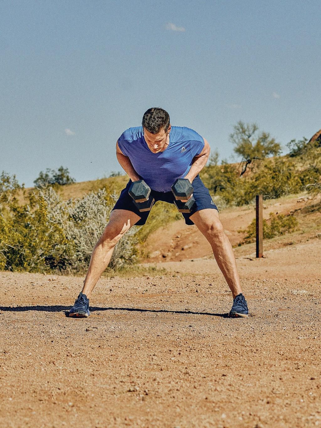 man in a straddle position, midway through rowing two dumbbells upwards