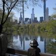 The Midtown Manhattan skyline is reflected in the water as people in masks walk along the lake in Central Park