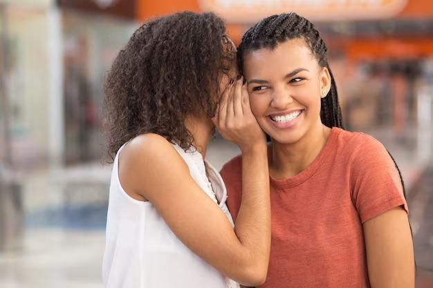 Black girl whispering secret to smiling girlfriend [Freepik]