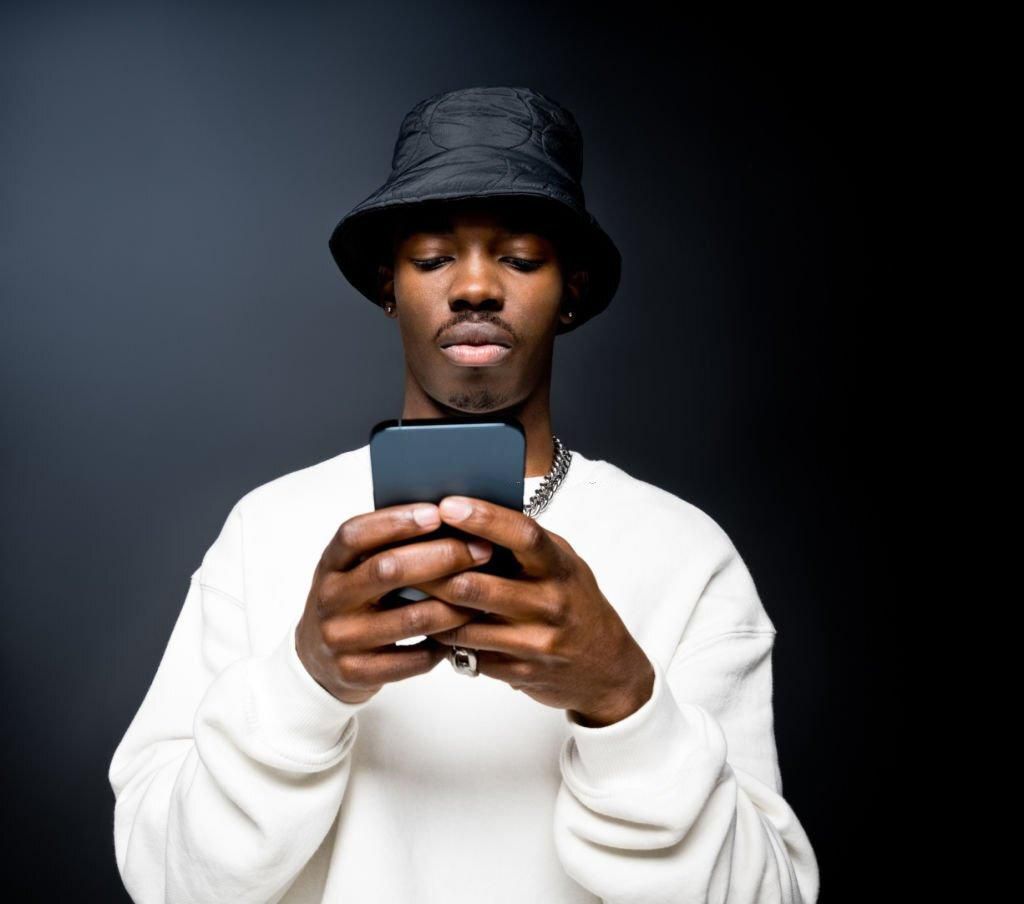 Portrait of handsome young man wearing white sweatshirt and black bucket hat, using mobile phone. Studio shot on black background.