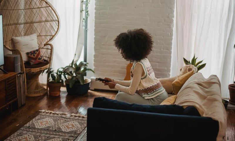 Focused young ethnic person with remote controller watching TV at home [Image credit: Andres Ayrton]