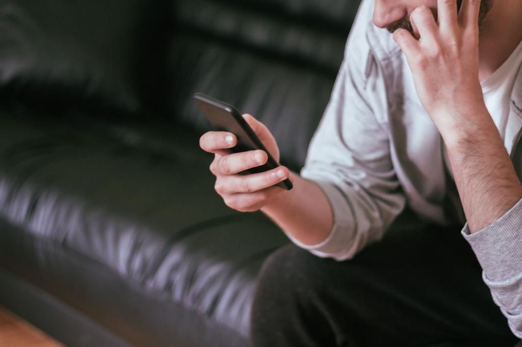 Serious bearded man is sitting on couch and typing on smartphone