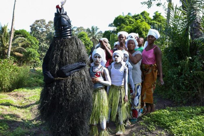 Young girls in white face paint take part in a procession to start their initiation into the Bondo secret society, alongside a figure called the Black Devil