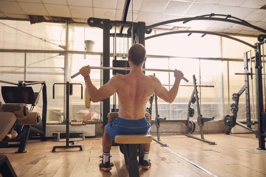 one young man, shirtless, back lat pulldown exercise, sitting multistationary machine in old beaten up gym interior. full length shot, rear view.
