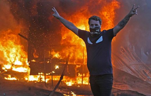 A protester flashes the V for victory sign as a riot police vehicle burns behind him during clashes amidst demonstrations against state corruption, failing public services, and unemployment in the Iraqi capital