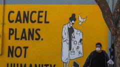 A man wearing gloves and a face mask walks by a mural reading "Cancel Plans Not Humanity" in Los Angeles, California
