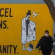 A man wearing gloves and a face mask walks by a mural reading "Cancel Plans Not Humanity" in Los Angeles, California