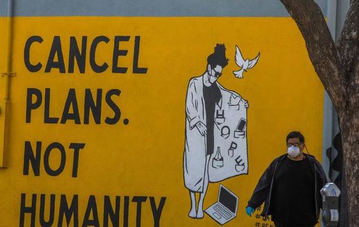 A man wearing gloves and a face mask walks by a mural reading "Cancel Plans Not Humanity" in Los Angeles, California