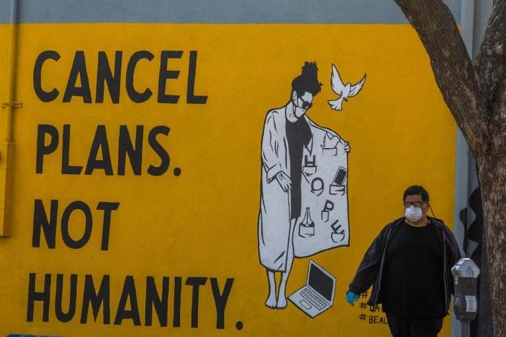 A man wearing gloves and a face mask walks by a mural reading "Cancel Plans Not Humanity" in Los Angeles, California