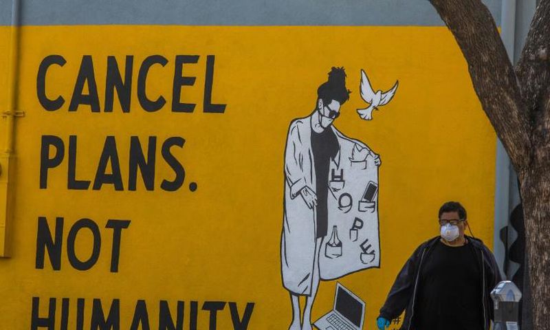 A man wearing gloves and a face mask walks by a mural reading "Cancel Plans Not Humanity" in Los Angeles, California