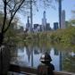 The Midtown Manhattan skyline is reflected in the water as people in masks walk along the lake in Central Park