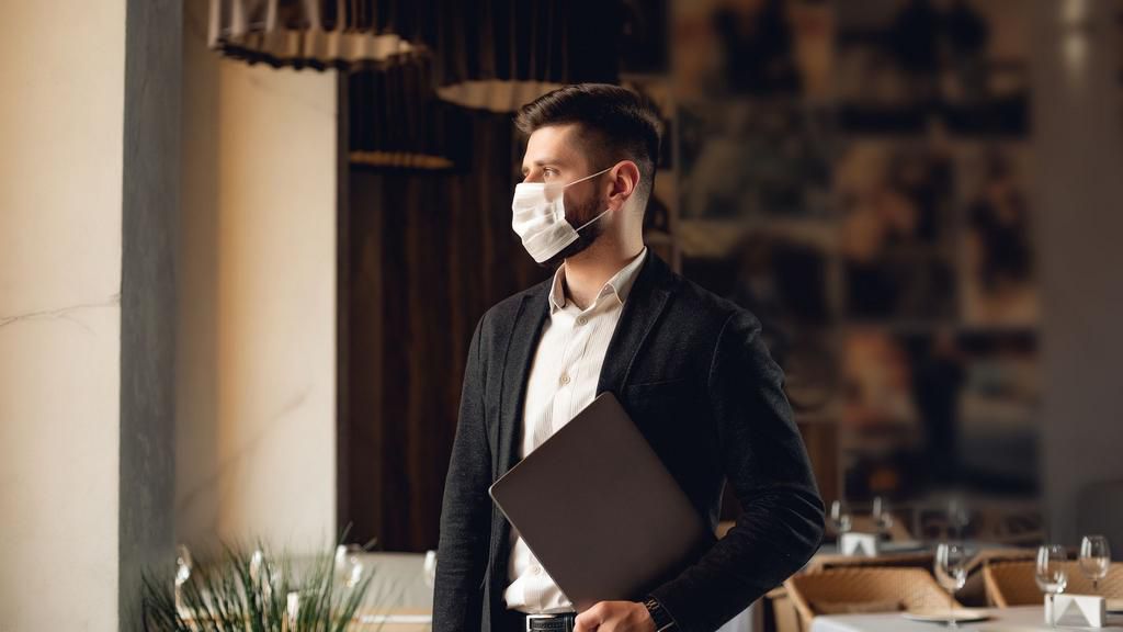 Handsome adult bearded man indoors in cafe. Lifestyle concept photo with copy space. Picture with gray laptop and protective mask on the face