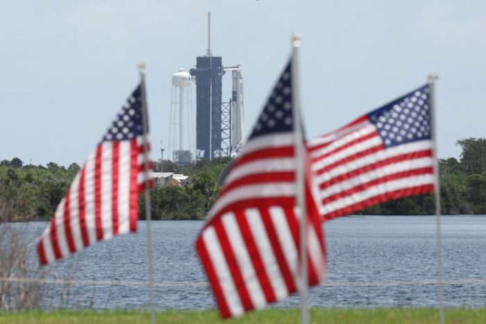 A SpaceX Falcon 9 rocket with the Crew Dragon spacecraft looms in the distance at launch complex 39A as American flags flutter in the wind, at the Kennedy Space Center in Florida on May 29