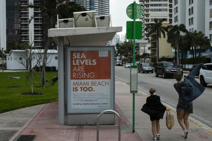 People walk past a billboard in Miami Beach discussing sea level rise: dozens of trucks have started dumping hundreds of thousands of tons of sand on Miami Beach as part of US government measures to protect Florida's tourist destinations against the ef...
