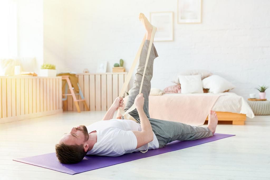 Calm relaxed handsome young man with beard lying on back and lifting leg while stretching it with yoga strap at home