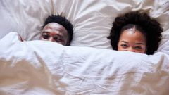 African couple peeking over their bedsheets while lying together in their bed