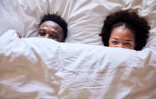 African couple peeking over their bedsheets while lying together in their bed