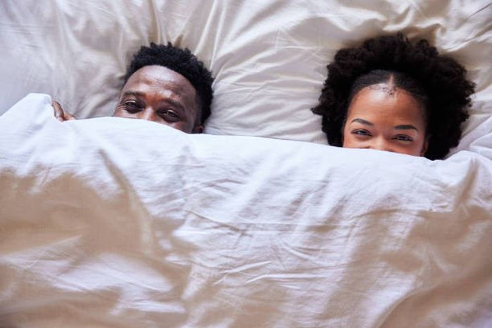 African couple peeking over their bedsheets while lying together in their bed