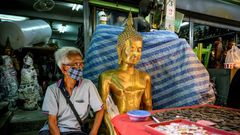 A vendor wearing a face mask against the spread of the coronavirus sits beside a Buddha statue as he waits for customers at an amulet market in Bangkok