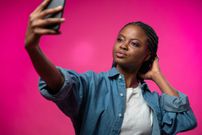 An attractive dark - blogger with afro braids in a jeans shirt and white t-shirt takes a selfie on her smart phone,