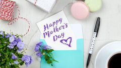 Happy Mother's Day card beside pen, macaroons, flowers, and gift box near a coffee cup uon a saucer [Photo: George Dolgikh]