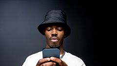 Portrait of handsome young man wearing white sweatshirt and black bucket hat, using mobile phone. Studio shot on black background.