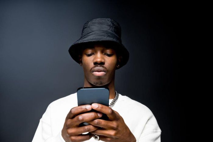 Portrait of handsome young man wearing white sweatshirt and black bucket hat, using mobile phone. Studio shot on black background.