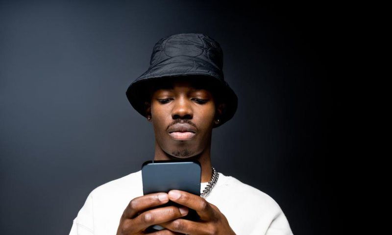 Portrait of handsome young man wearing white sweatshirt and black bucket hat, using mobile phone. Studio shot on black background.