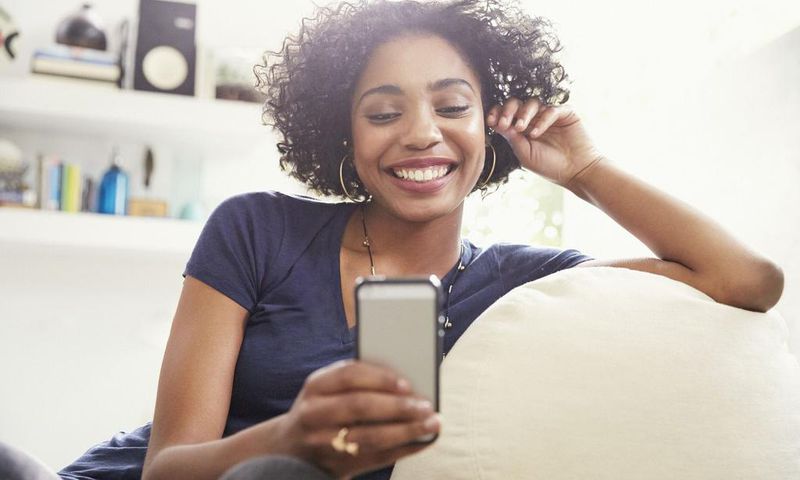 Smiling African American woman texting with cell phone on living room sofa